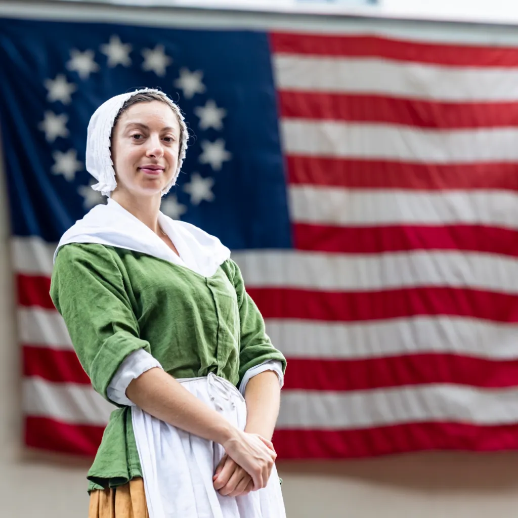A History Maker portraying Betsy Ross stands in front of a large version of the early US flag in the courtyard of the Betsy Ross House