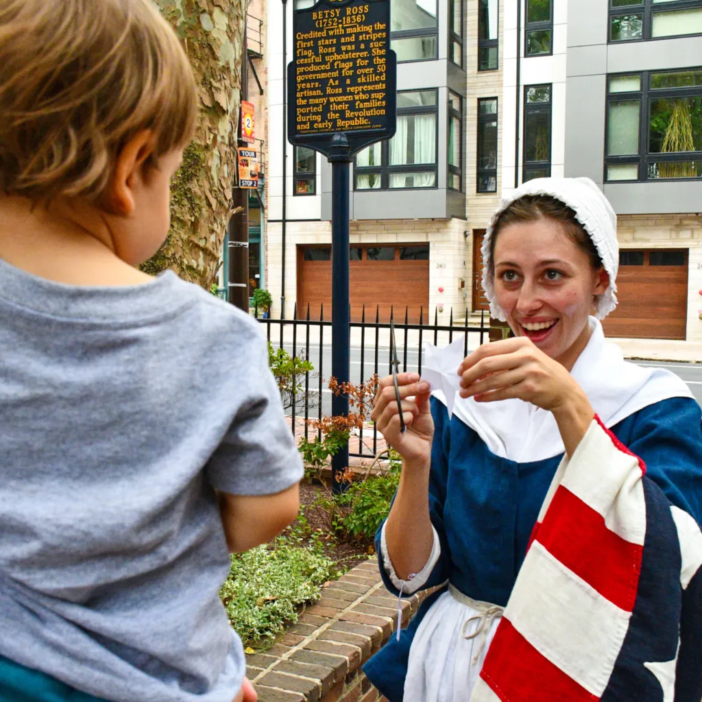 A History Maker portraying Betsy Ross cuts a star out of paper and shows it to a child