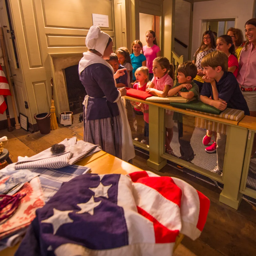 Una creadora de historia que representa a Betsy Ross habla con un grupo de niños y adultos en el taller de tapicería durante un recorrido por la casa de Betsy Ross.