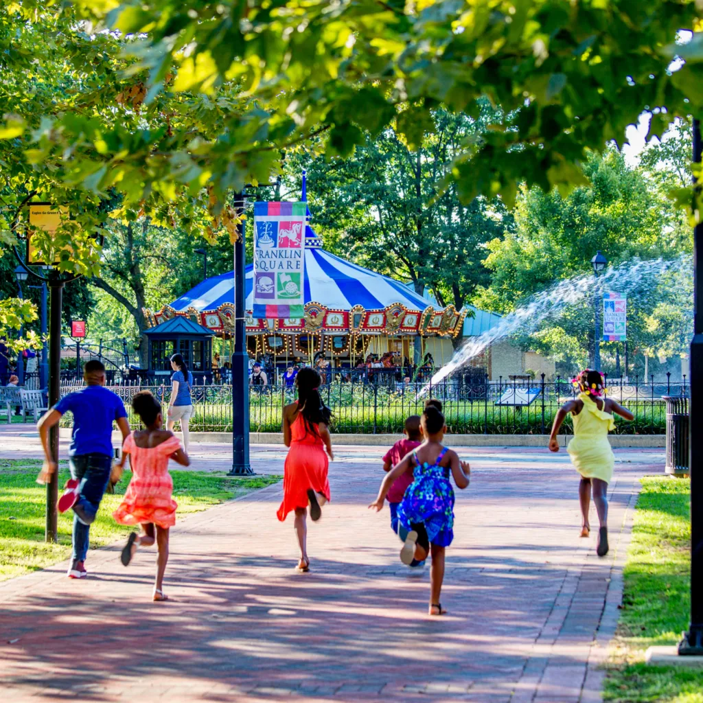 A group of children run towards the center of Franklin Square with the fountain and carousel in the background
