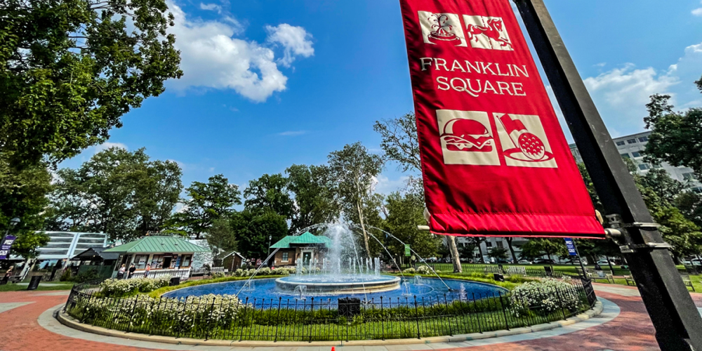 A wide angle view of the Franklin Square park fountain with a sign in the foreground that says 