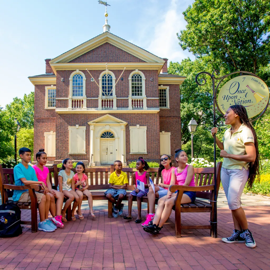 Children sit on a bench at Carpenters Hall while a Once Upon A Nation Story Teller shares a story