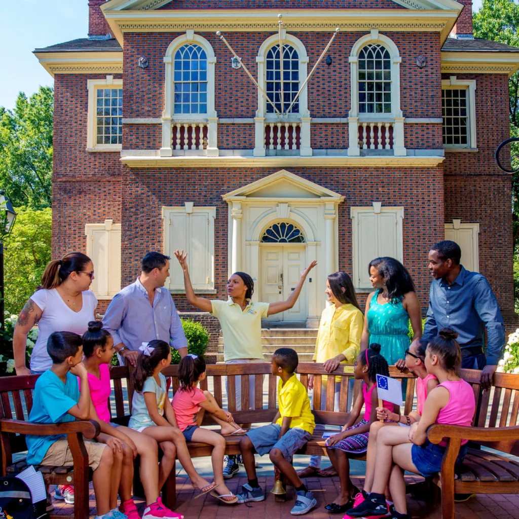 Adults and children gather around a storytelling bench outside Independence Hall to listen to a Storyteller tell a short story