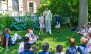 A group of children sit in a circle in the grass as they listen to a Storyteller tell a story on the Story Stroll tour
