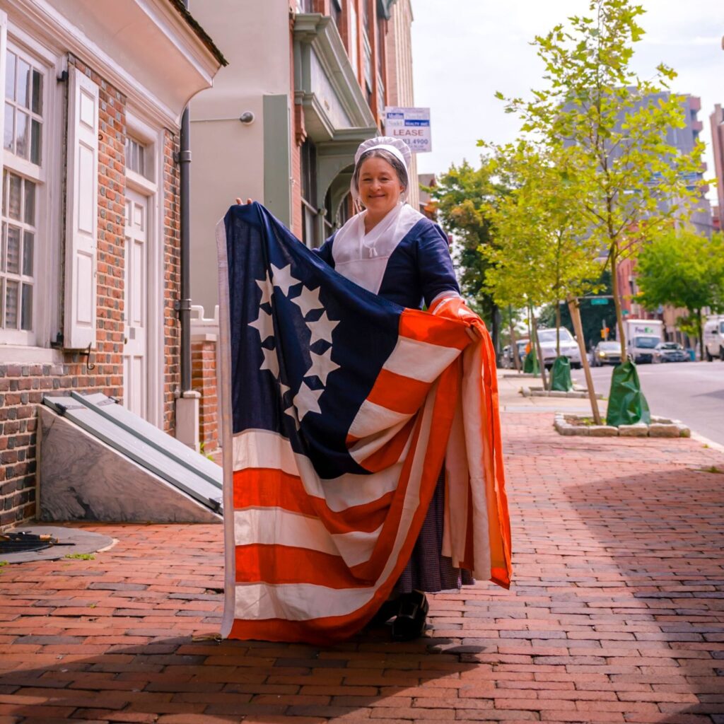 Betsy Ross holding the 13-starred flag outside of the Betsy Ross House