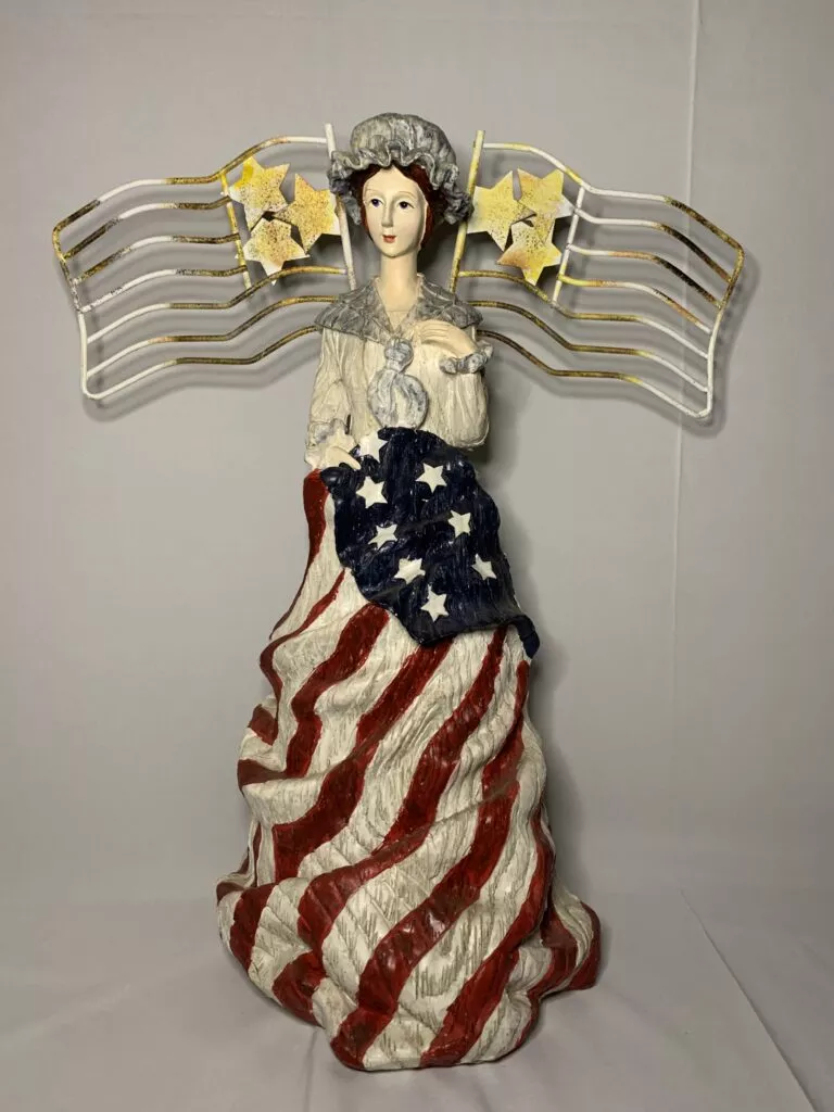 An angelic figure modeled after Betsy Ross holding two flags across her chest made by the Bayberry Family Collection.