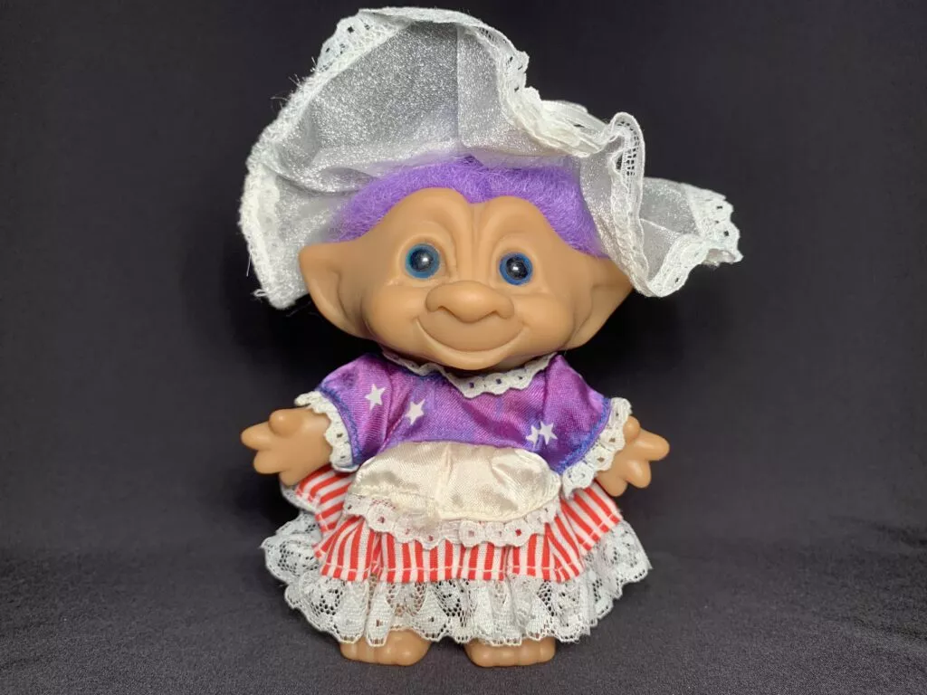 Troll Doll inspired by Betsy Ross. the doll has purple hair and is wearing red white and blue colonial garb.