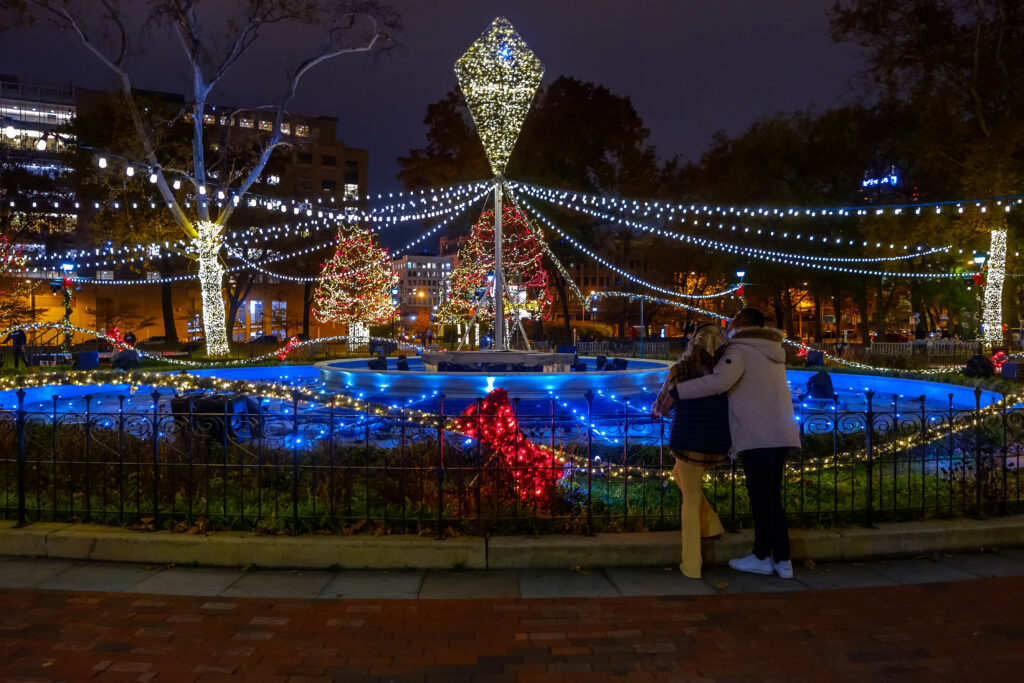 Photo of a couple embracing next to the Rendell Family Fountain during the Electrical Spectacle Light Show presented by PECO during Winter in Franklin Square.
